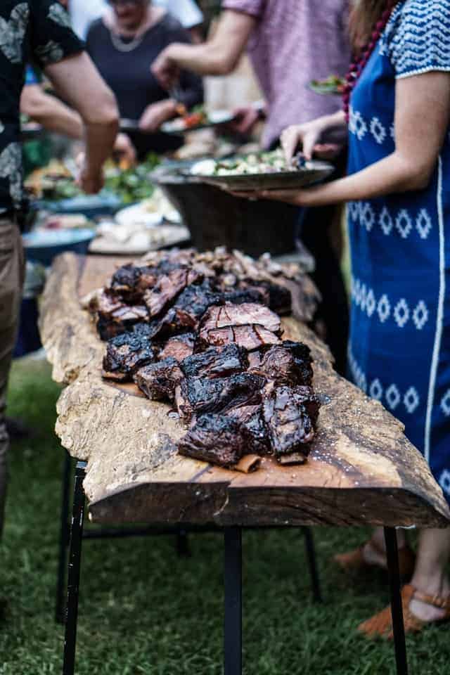 Schwarz gegrilltes Fleisch auf Holztisch mit Festgesellschaft im Hintergrund
