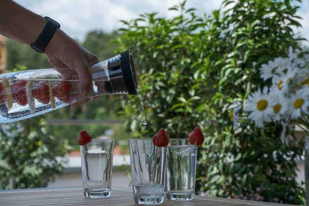 Männerhand schenkt Wasser aus Karaffe in drei Gläser mit dekorativen Erdbeeren, Im Hintergrund ist ein Garten zu sehen