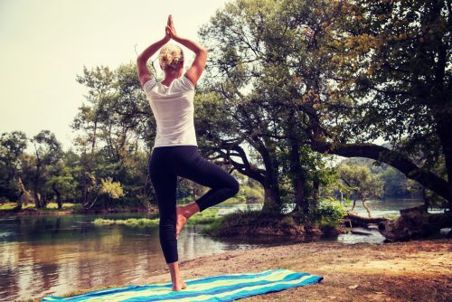 Junge Frau in Yoga-Haltung an Seeufer mit Bäumen im Hintergrund