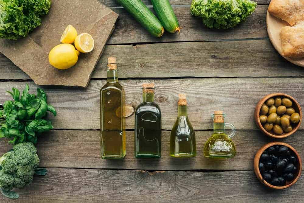Auf Holztisch arrangierte Ölflaschen mit grünen und schwarzen Oliven in Tonschälchen und Gemüse im Hintergrund arrangiert