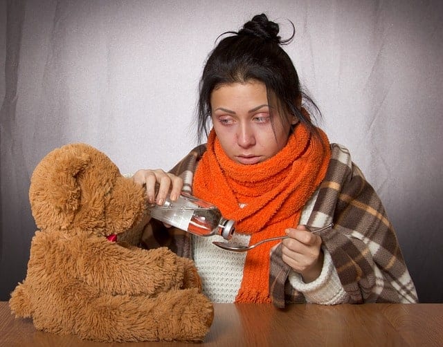Erkältete Frau mit roten Augen, Schal, Hustensaft und tröstendem Teddybär