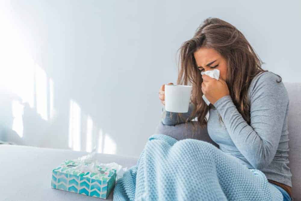 Frau mit Erkältung und einer Tasse Tee in der Hand benutzt ein Taschentuch auf heller Couch
