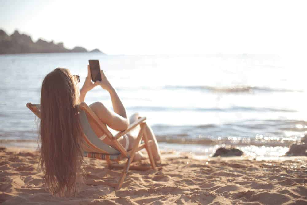 Gefahren von UV Strahlung - junge Frau sitzt mit Handy im Liegestuhl in der Sonne am Meer