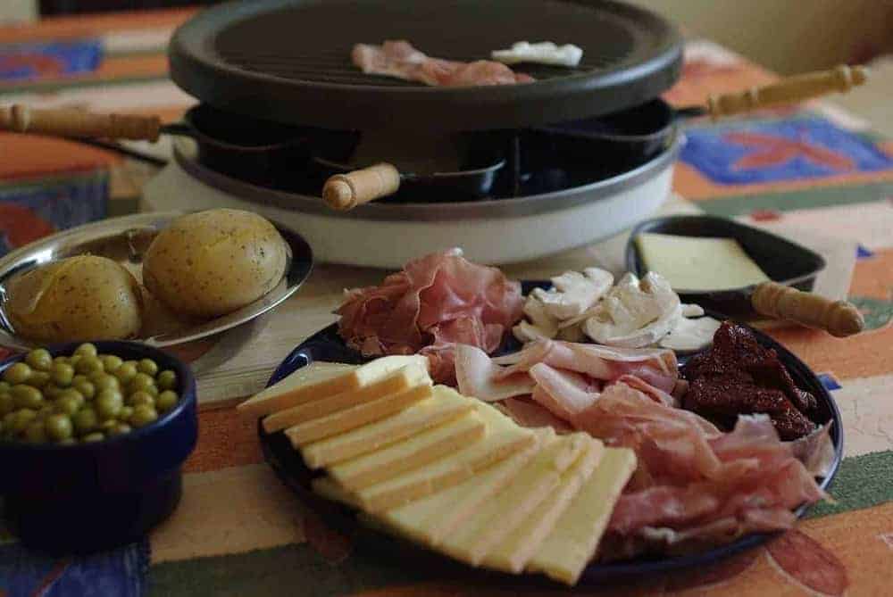 Raclette-Grill mit Schinken-Käse-Platte, Kartoffeln und grünen Erbsen auf Esstisch
