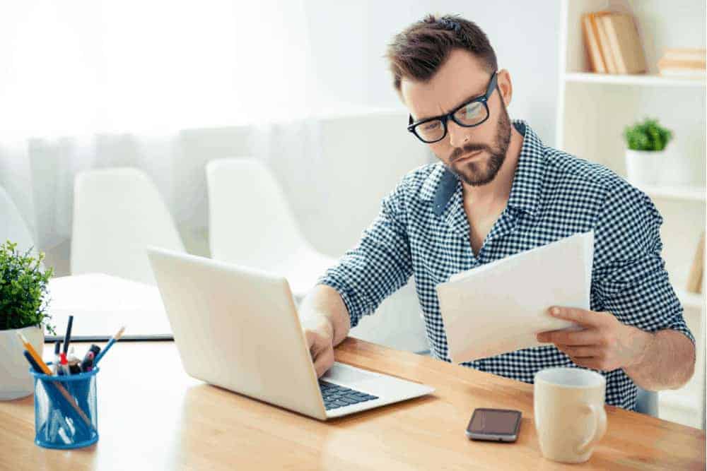 Junger Mann mit Brille sitzt vor Laptop mit Tasse Kaffee