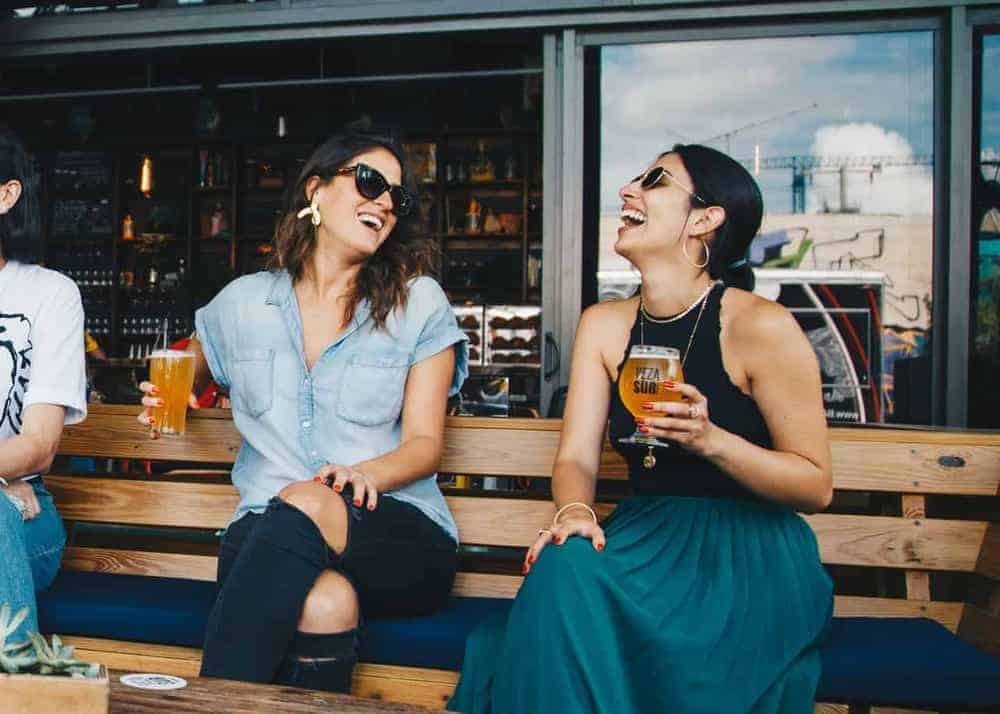 Zwei junge Frauen mit Sonnenbrille und Glas Bier in der Hand sitzen vor einem Restaurant