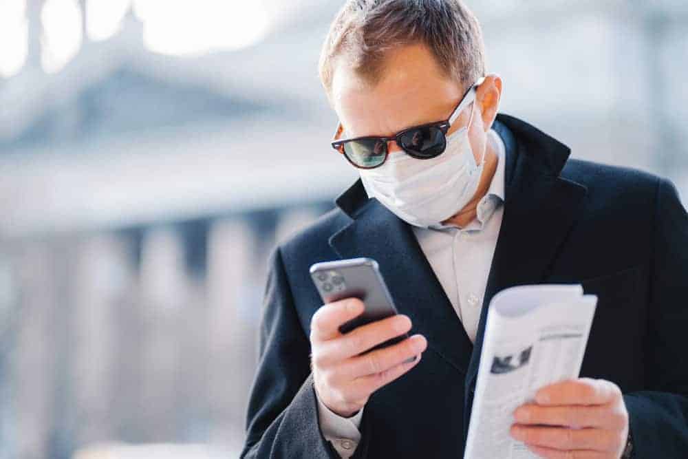 Manager mit Mundschutz und Sonnenbrille, Trenchcoat und Smartphone in der Stadt will Mundschutz kaufen