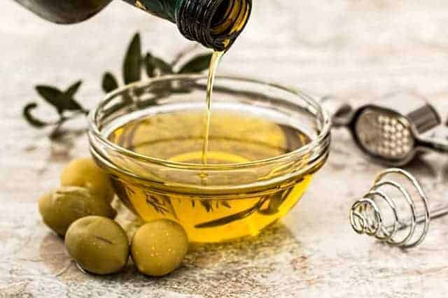Olivenöl in Glasschale neben frischen grünen Oliven und Olivenzweig im Hintergrund