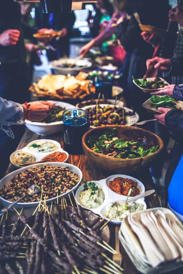 Festtafel mit Fleisch-Spießen, Brot, Salat, Oliven; im Hintergrund Gäste, die sich bedienen