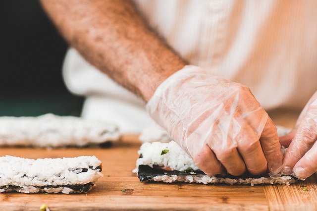 Mann mit Handschuh rollt Klebreis zu Sushi-Rolle auf Holztisch