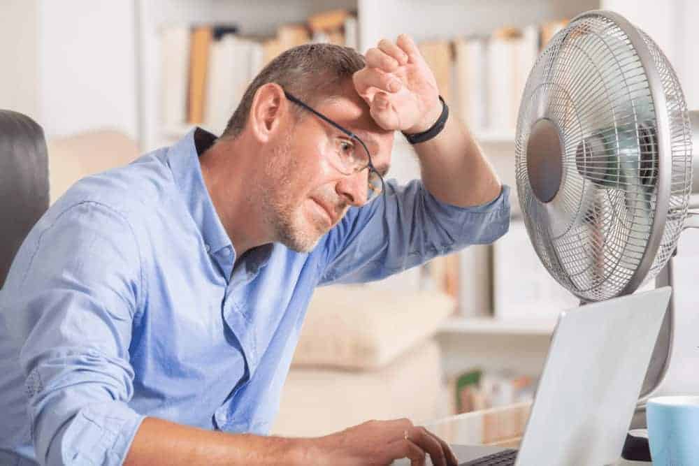 Tipps gegen die Sommerhitze Mann im Büro am Schwitzen mit Ventilator und Schweiß auf der Stirn
