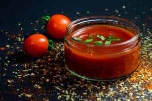 Tomatensauce in Glas vor Tomaten auf schwarzem Hintergrund mit Gewürzen