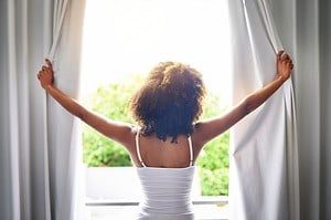 Junge Frau öffnet die Vorhänge eines Fensters, sodass Sonne hereinscheint